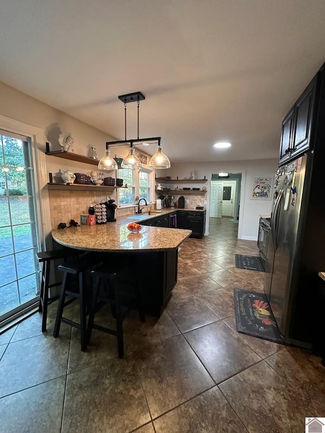 kitchen with dark cabinets, hanging light fixtures, open shelves, stainless steel fridge, and a kitchen bar