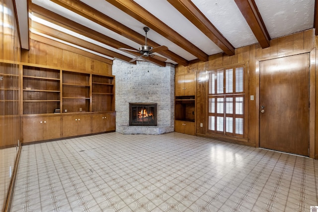 unfurnished living room featuring ceiling fan, a stone fireplace, wood walls, and beam ceiling