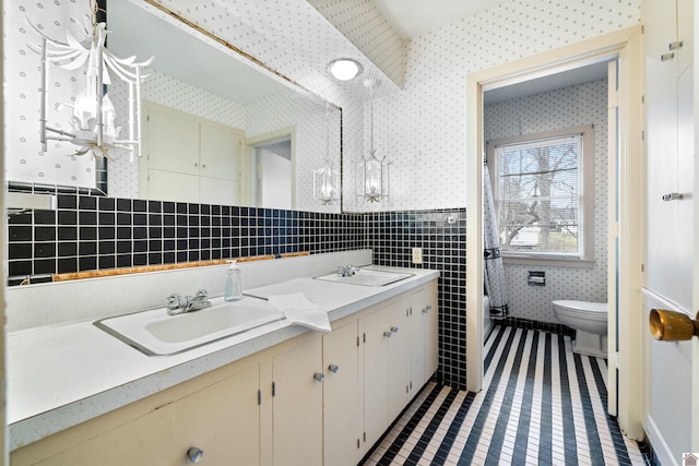 bathroom featuring double vanity, wallpapered walls, a sink, and wainscoting