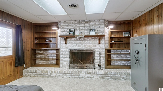 unfurnished living room featuring carpet floors, built in shelves, visible vents, a brick fireplace, and wooden walls