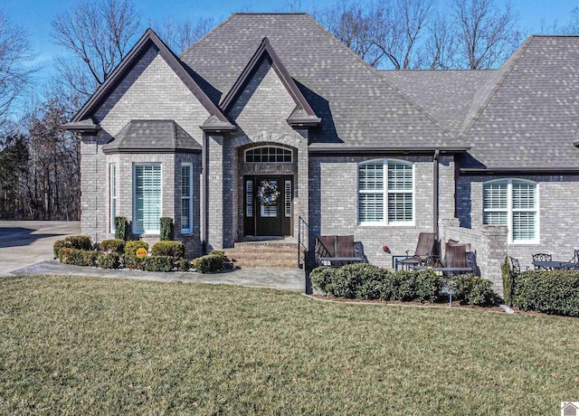 french country style house featuring roof with shingles, a front yard, and brick siding