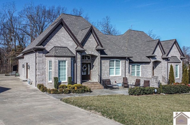 french country home featuring brick siding, roof with shingles, a front lawn, and a patio