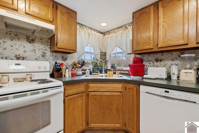 kitchen with under cabinet range hood, white appliances, a sink, dark countertops, and wallpapered walls