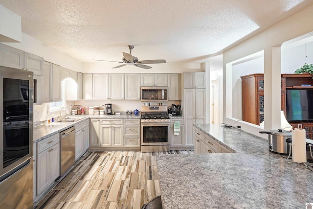 kitchen with stainless steel appliances, a sink, a ceiling fan, light stone countertops, and light wood finished floors