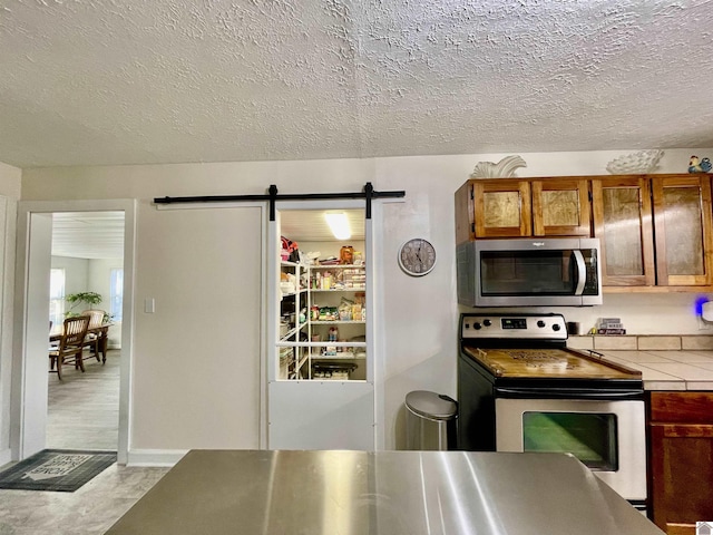 kitchen featuring tile countertops, a textured ceiling, a barn door, appliances with stainless steel finishes, and brown cabinets