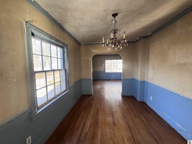 unfurnished dining area featuring crown molding, dark wood-type flooring, a wainscoted wall, arched walkways, and a notable chandelier