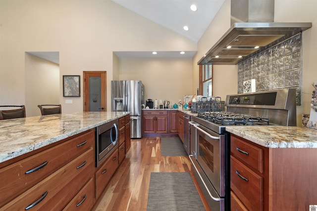 kitchen with light stone countertops, vaulted ceiling, light wood-style flooring, stainless steel appliances, and wall chimney exhaust hood