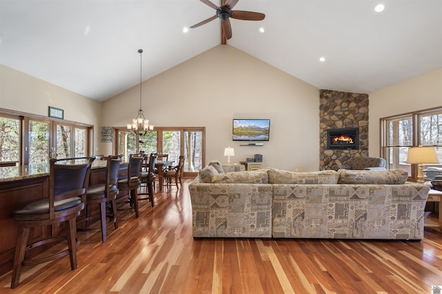 living room featuring high vaulted ceiling, a stone fireplace, wood finished floors, and ceiling fan with notable chandelier