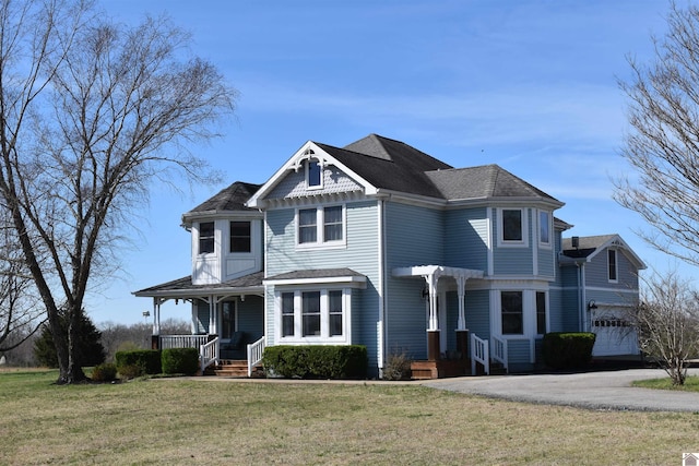 victorian-style house with a front yard, covered porch, a shingled roof, a garage, and aphalt driveway
