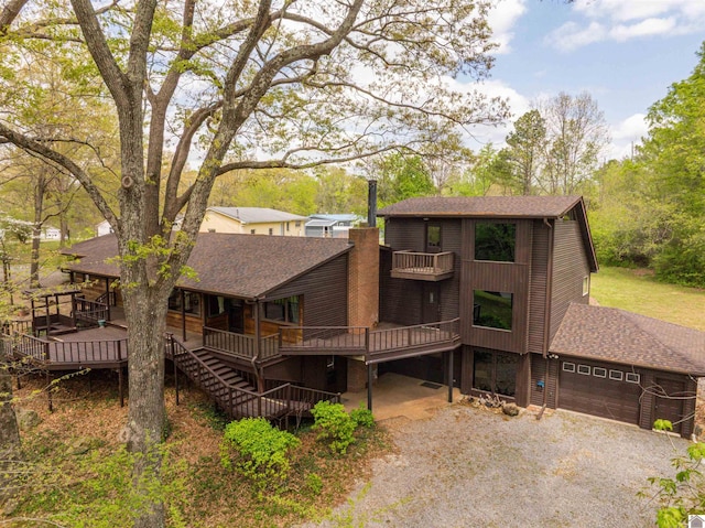 view of front of house with a wooden deck, an attached garage, a shingled roof, a balcony, and gravel driveway