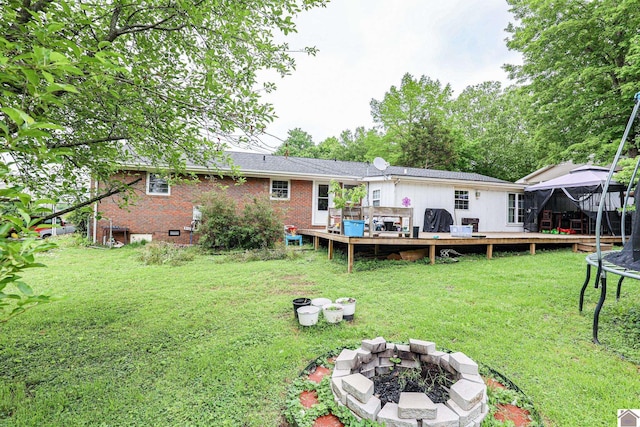 rear view of house with a deck, a trampoline, a fire pit, brick siding, and a lawn