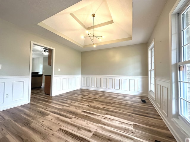empty room featuring a tray ceiling, a chandelier, wood finished floors, a wainscoted wall, and a decorative wall