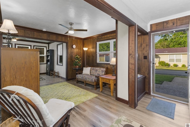 living room featuring crown molding, wooden walls, light wood finished floors, baseboards, and ceiling fan