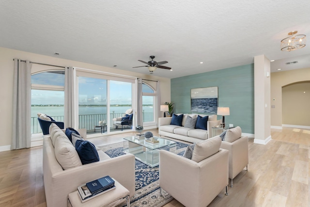 living room with ceiling fan with notable chandelier, a water view, light hardwood / wood-style floors, and a textured ceiling