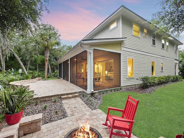 back house at dusk with a sunroom, ceiling fan, a yard, a patio, and an outdoor fire pit