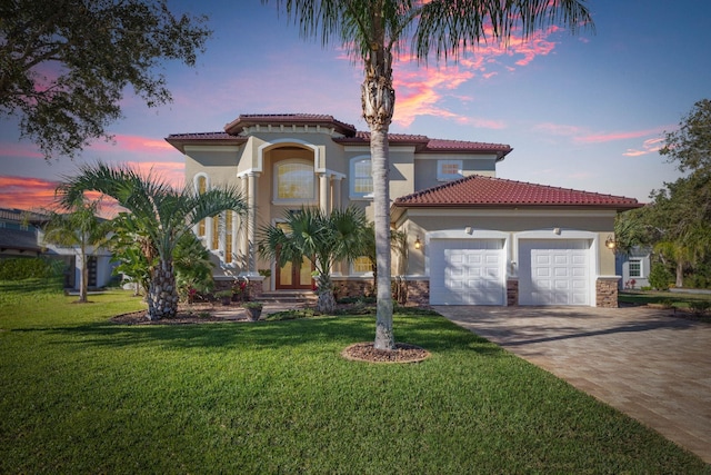 mediterranean / spanish home featuring decorative driveway, stucco siding, a lawn, a garage, and a tiled roof