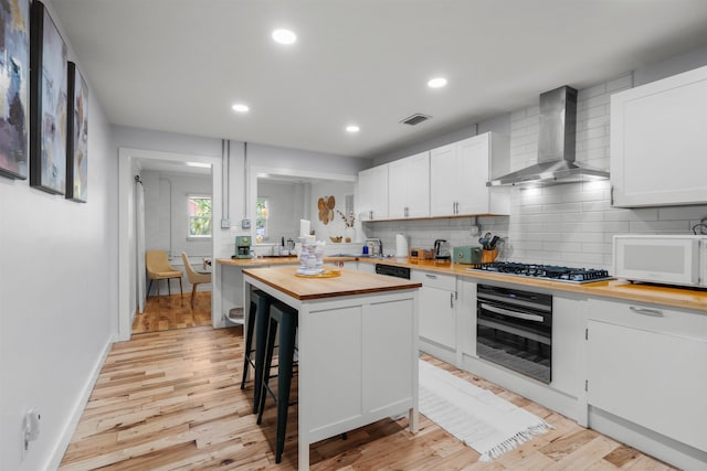 kitchen with wall chimney range hood, light wood finished floors, wooden counters, visible vents, and black appliances