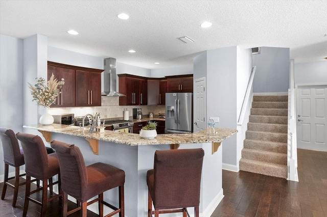 kitchen featuring wall chimney range hood, dark wood-type flooring, appliances with stainless steel finishes, backsplash, and kitchen peninsula