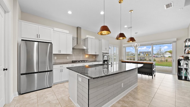 kitchen with wall chimney range hood, decorative light fixtures, a kitchen island with sink, white cabinets, and appliances with stainless steel finishes