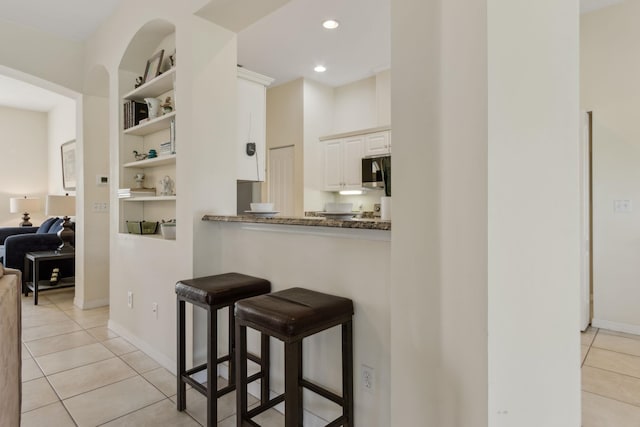 kitchen featuring light tile patterned floors, stainless steel microwave, a breakfast bar area, built in shelves, and white cabinetry