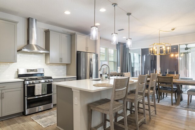 kitchen with wall chimney range hood, appliances with stainless steel finishes, gray cabinetry, a center island with sink, and decorative light fixtures