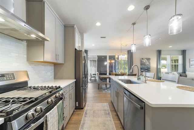 kitchen featuring gray cabinets, appliances with stainless steel finishes, pendant lighting, a center island with sink, and wall chimney exhaust hood