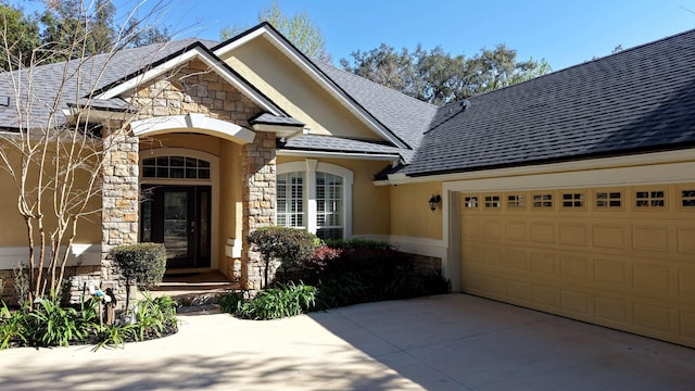 view of front facade featuring driveway, an attached garage, a shingled roof, stucco siding, and stone siding