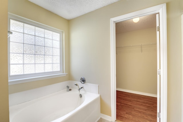 full bathroom with wood finished floors, baseboards, a textured ceiling, a walk in closet, and a garden tub