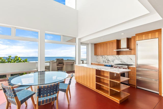 kitchen featuring stainless steel built in fridge, a water view, wall chimney exhaust hood, decorative backsplash, and a towering ceiling