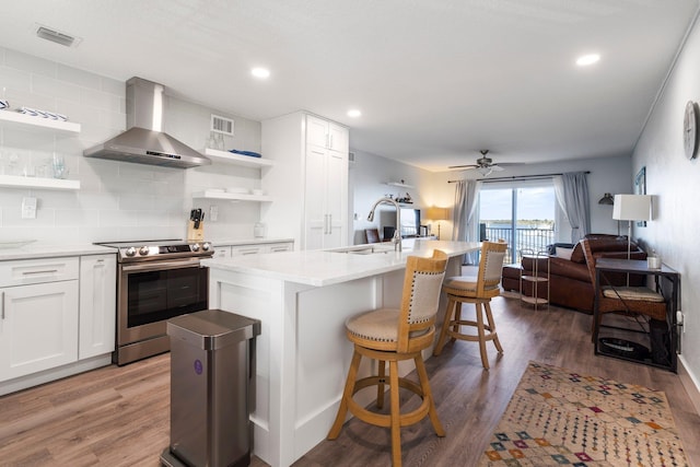 kitchen with stainless steel range with electric cooktop, wall chimney exhaust hood, white cabinets, and a center island with sink