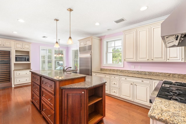 kitchen featuring crown molding, exhaust hood, built in appliances, hanging light fixtures, and an island with sink