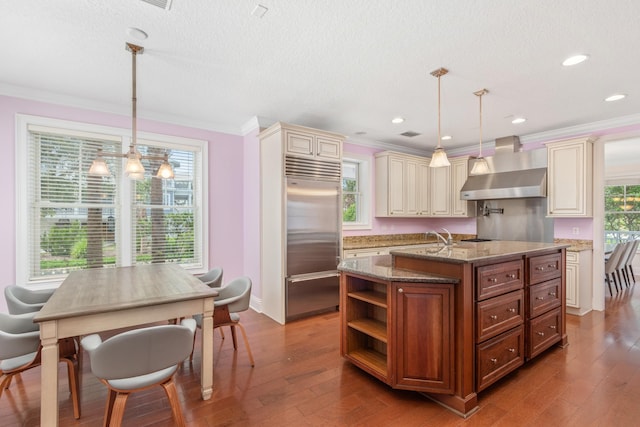 kitchen with pendant lighting, stainless steel built in fridge, wall chimney range hood, and an island with sink