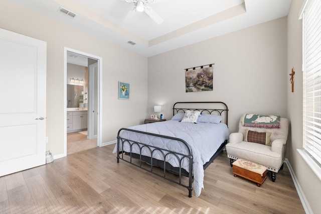 bedroom with ensuite bathroom, a raised ceiling, ceiling fan, and light wood-type flooring