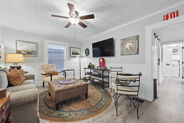 living area featuring an ornate ceiling, crown molding, light wood-style flooring, wainscoting, and ceiling fan