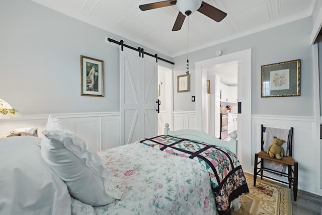 bedroom with a barn door, a wainscoted wall, wood finished floors, ornamental molding, and an ornate ceiling