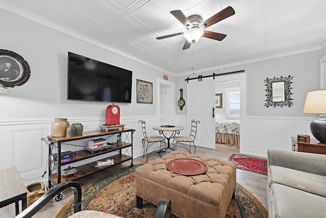 living room with a barn door, a ceiling fan, a wainscoted wall, an ornate ceiling, and wood finished floors