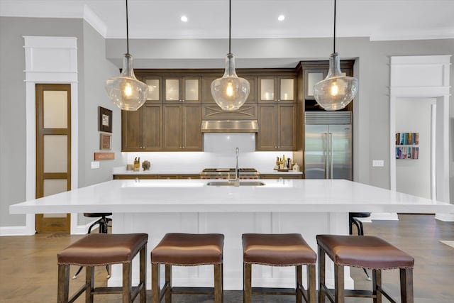 kitchen with glass insert cabinets, extractor fan, an island with sink, stainless steel built in fridge, and ornamental molding