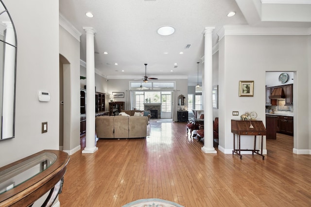 living room featuring ornate columns, ceiling fan, a stone fireplace, light hardwood / wood-style flooring, and crown molding