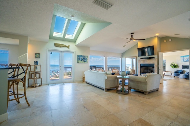 living room with a tile fireplace, ceiling fan, french doors, a textured ceiling, and vaulted ceiling with skylight