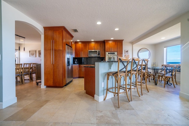 kitchen featuring a breakfast bar, dark stone countertops, a textured ceiling, an island with sink, and appliances with stainless steel finishes