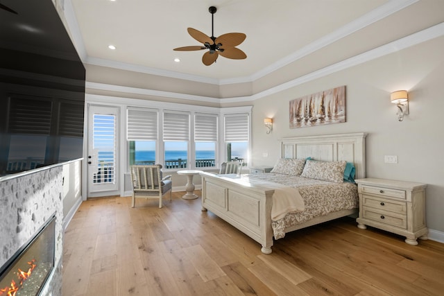 bedroom with crown molding, access to exterior, a water view, light hardwood / wood-style floors, and a stone fireplace