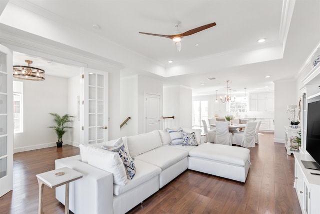 living room with ceiling fan with notable chandelier, a raised ceiling, dark wood-type flooring, and crown molding