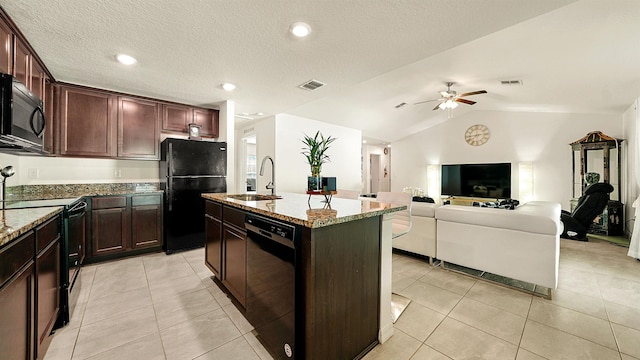 kitchen featuring lofted ceiling, black appliances, a center island with sink, sink, and light stone countertops