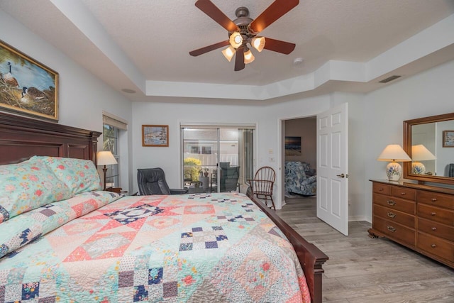 bedroom with access to outside, ceiling fan, a textured ceiling, a tray ceiling, and light hardwood / wood-style floors