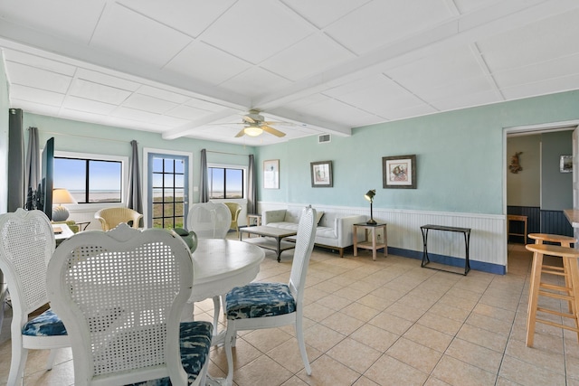 dining room with a wainscoted wall, beam ceiling, light tile patterned flooring, visible vents, and a ceiling fan