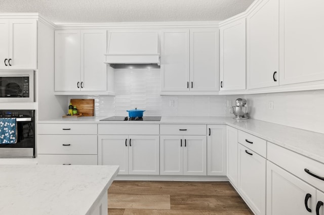 kitchen featuring black appliances, premium range hood, white cabinets, light wood-style flooring, and tasteful backsplash
