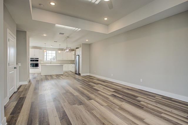 unfurnished living room with ceiling fan, sink, light wood-type flooring, and a tray ceiling