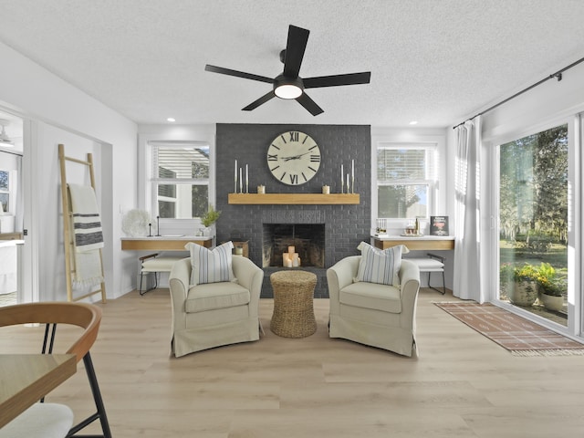 living room featuring light wood-type flooring, a brick fireplace, a textured ceiling, and ceiling fan