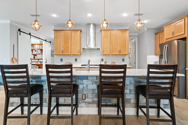 kitchen with wall chimney range hood, light brown cabinets, light countertops, decorative backsplash, and stainless steel refrigerator with ice dispenser