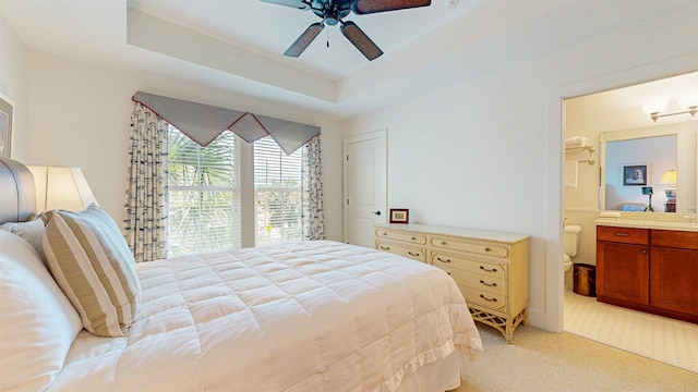 bedroom with light colored carpet, connected bathroom, a tray ceiling, and a ceiling fan